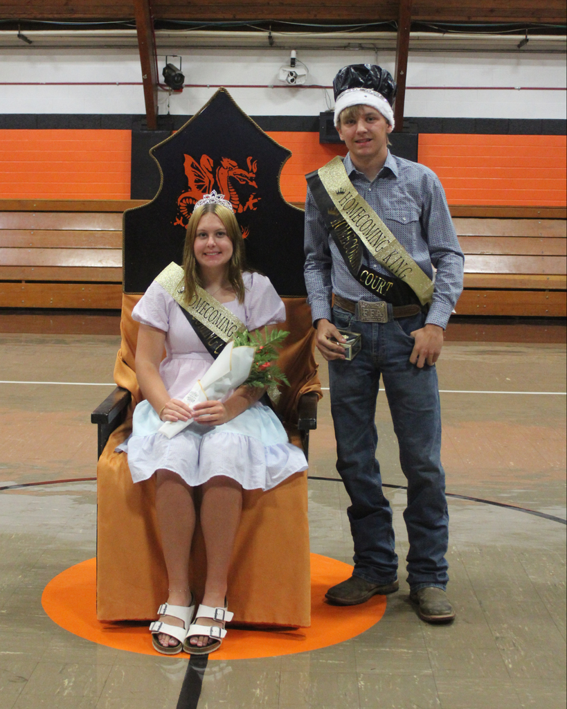 A teenage girl wearing a tiara, sitting in a chair while a teenage boy wearing a crown stands beside her
