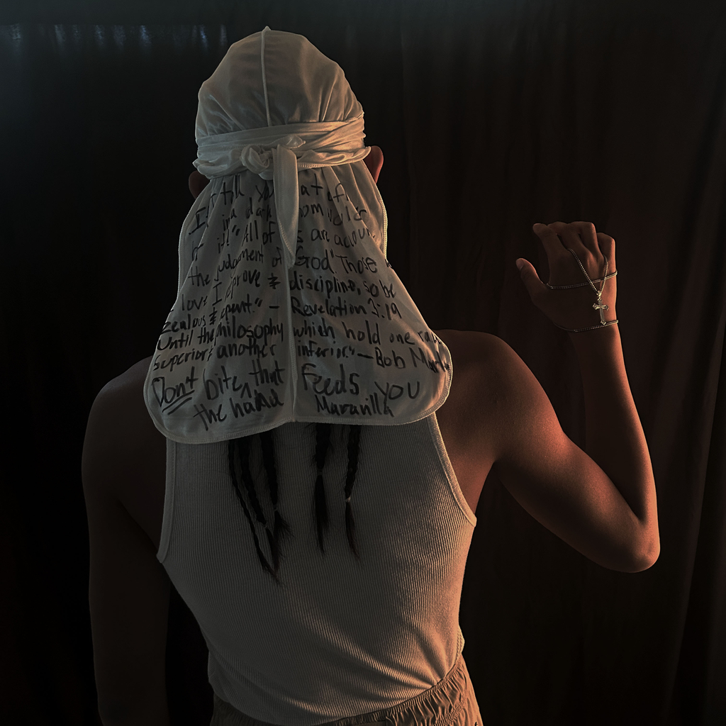 Person holding hand up with a cross and a head covering  with words that are slightly hard to read on it. 