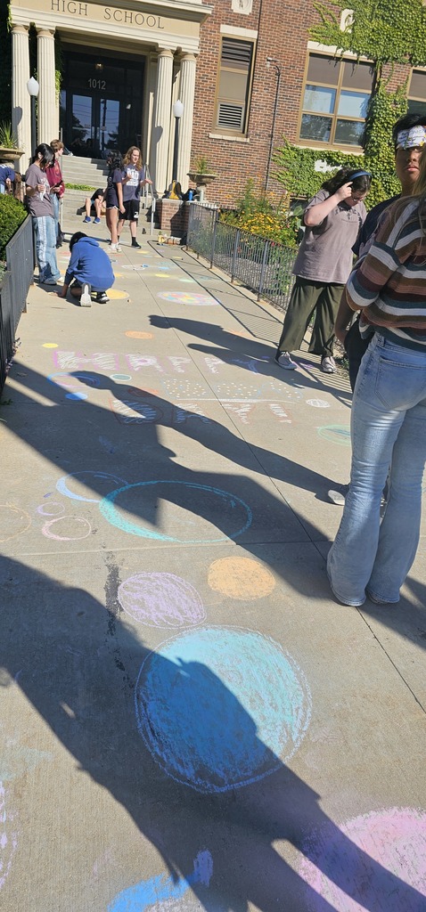 Students by the sidewalk filled with chalk. The sidewalk says, "Happy dot day!'