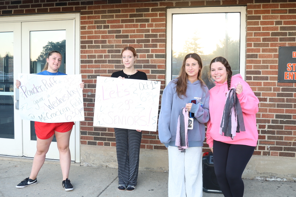 Students holding signs that say Powder Puff Let's go Seniors October 1. other students holding pom poms 