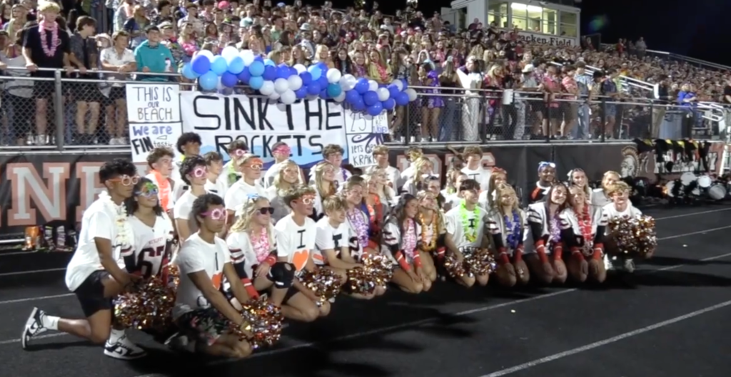 Students dressed up in sunglasses and Hawaiian leis with pom poms in front of balloons at the football game that says "sink the rocket"