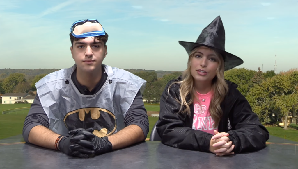 Two students sitting at a news desk. One in a Batman costume. The other in a witch costume