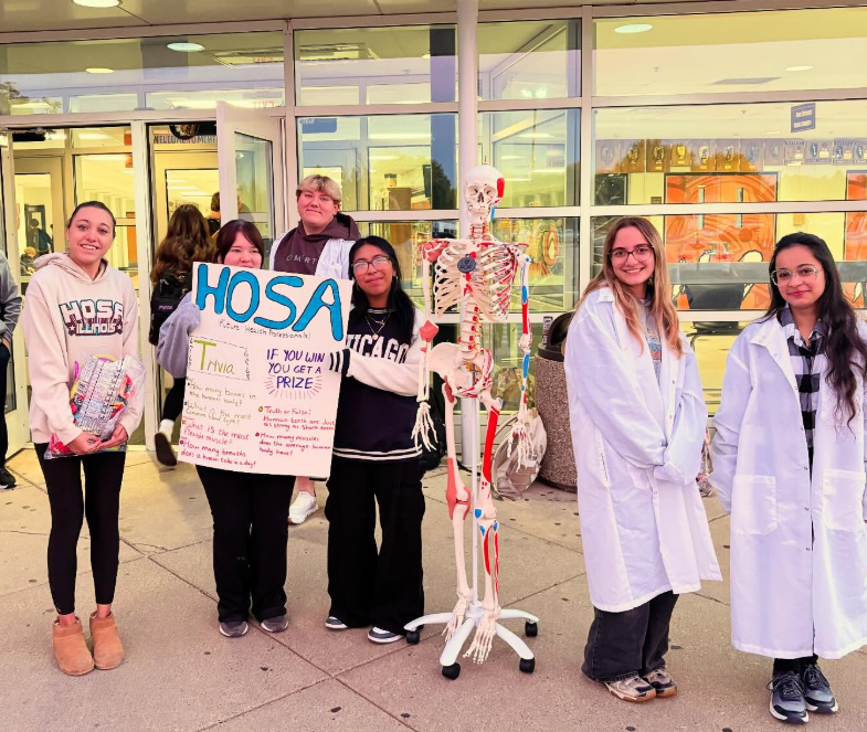 Students posing next to a HOSA Sign and a skeleton display. Two students in lab coats