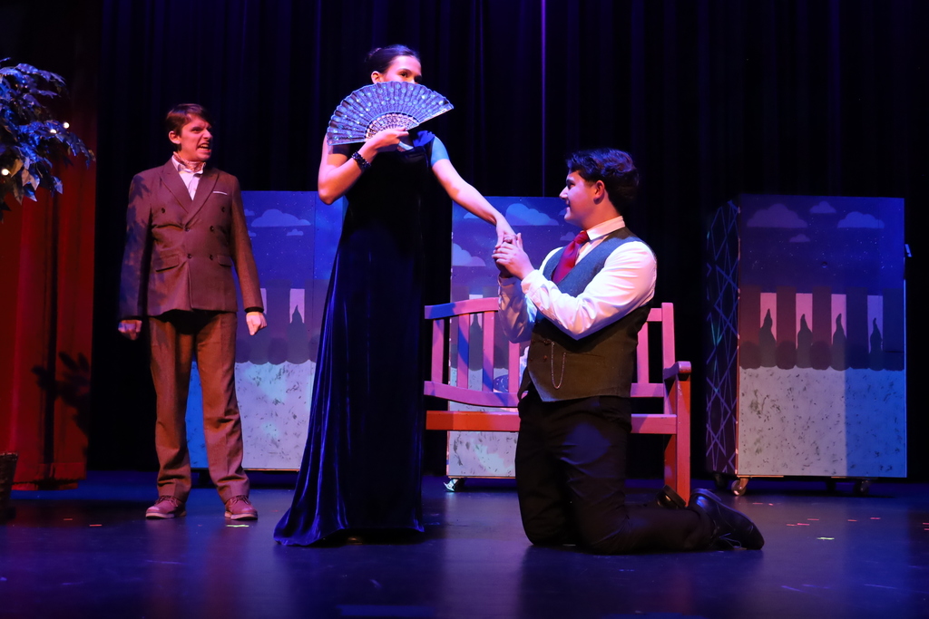 Student with dress and fan over her face, other character on knees holding her hand and other character in the background looking angry