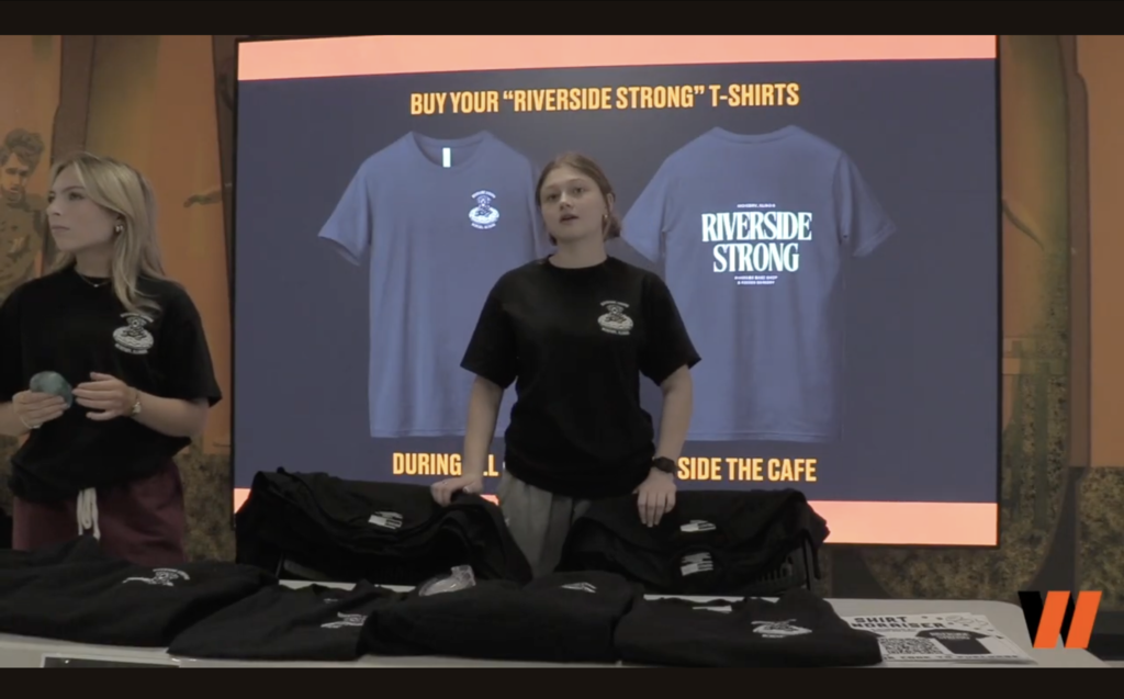 Student with t-shirts on a table. In the back, "Buy your Riverside Strong T-shirts"