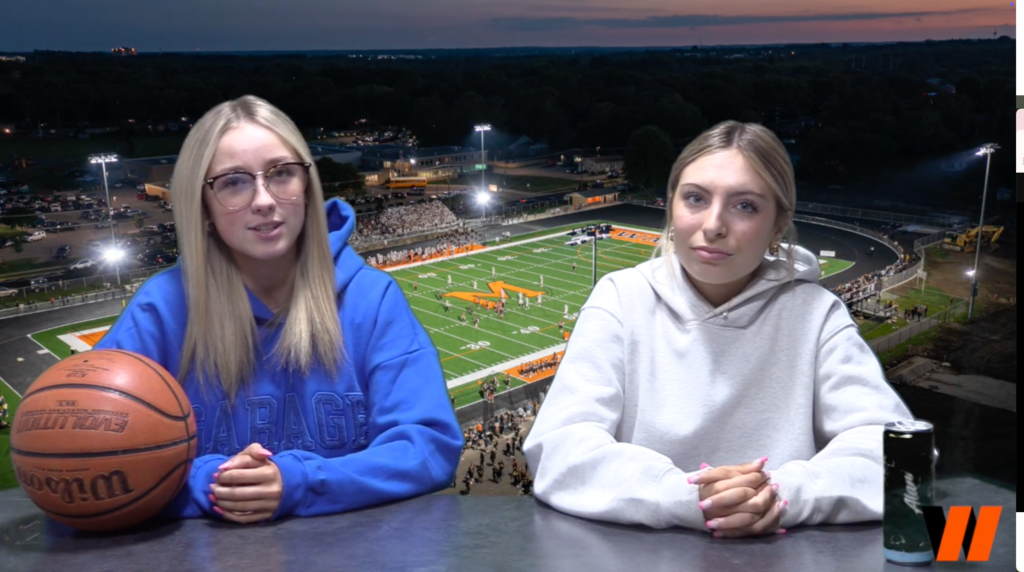 Two students in front of football field backdrop with a basketball at a desk 