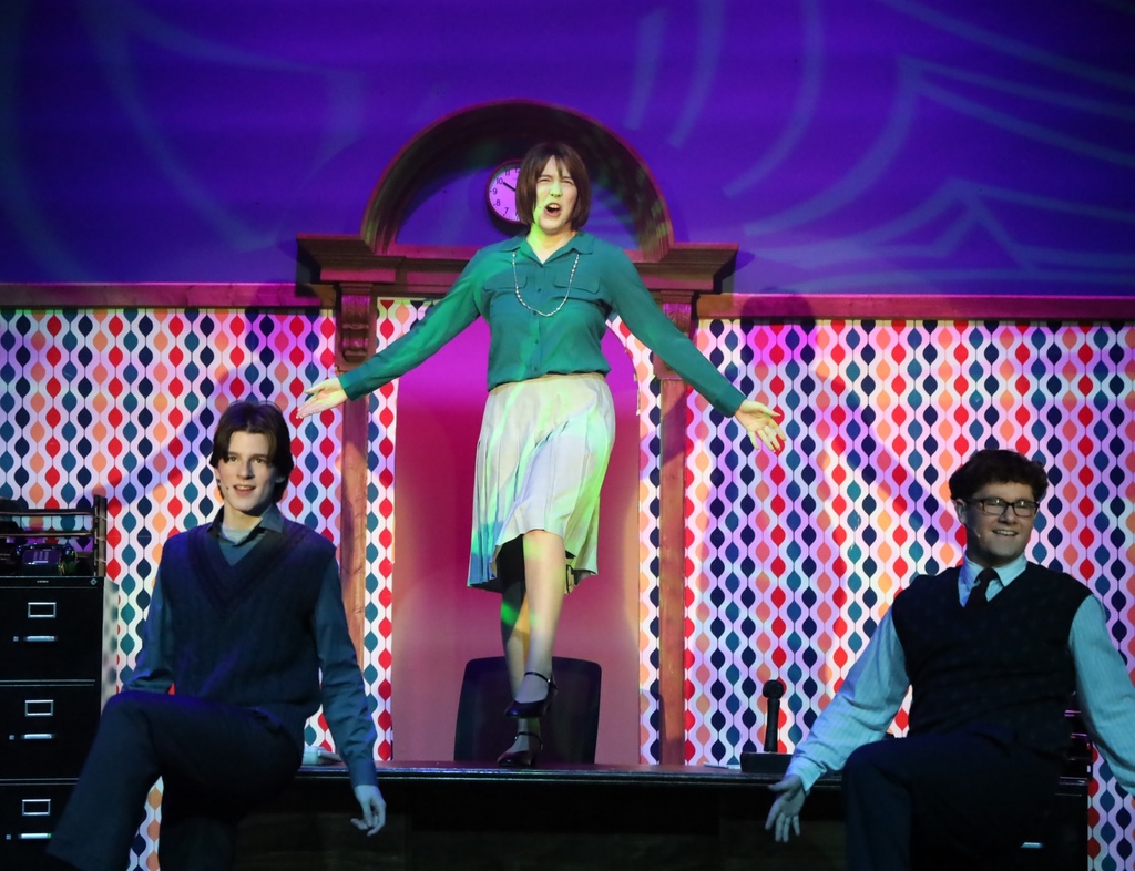 Women standing on desk with two men standing beside her on the ground.