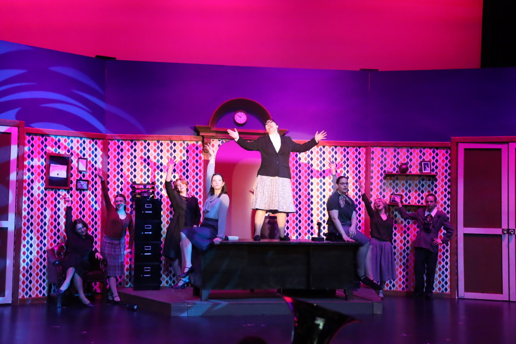 Female character standing on desk singing with other office girls around her