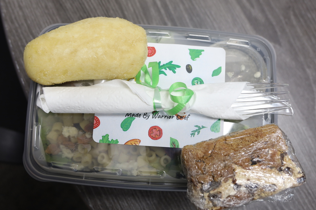 Container of salad with a label that says made by Warrior Chef. A roll, silverware and brownie 