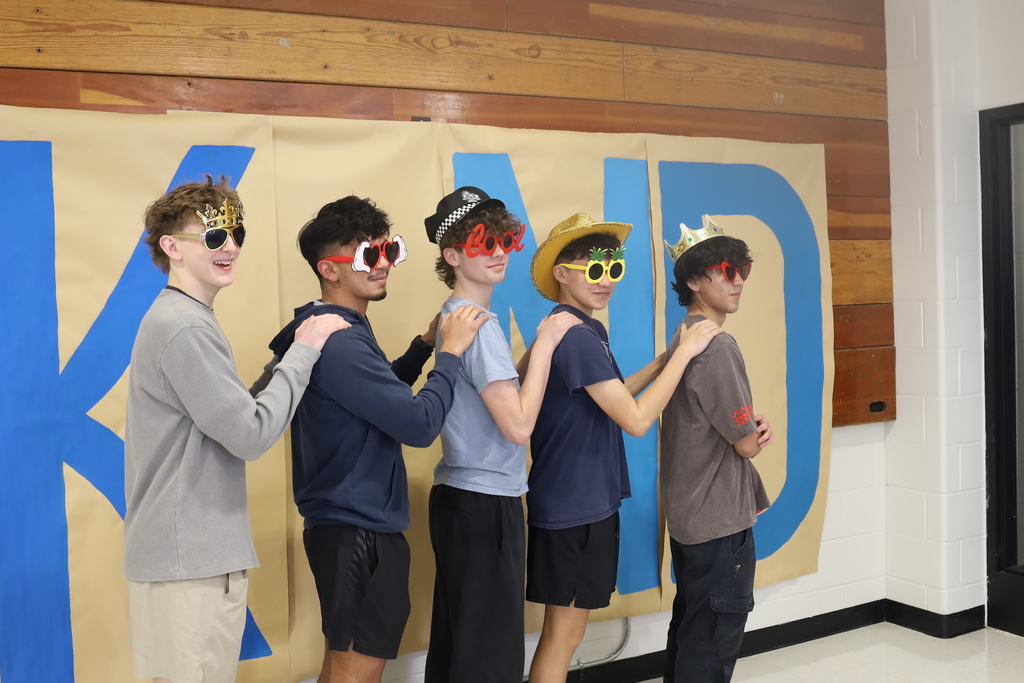 Four students standing in a conga line in front of Kind wall with sunglasses and fun hats