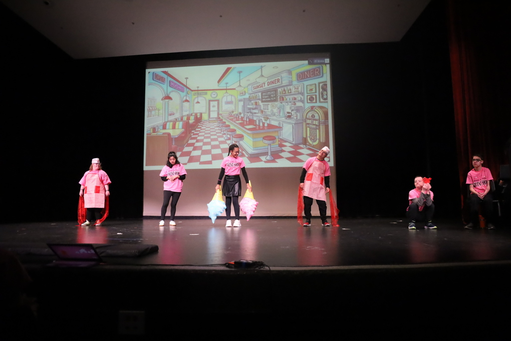 Students on stage dancing, one holding two inflatable ice creams, two dressed like classic food workers, diner cartoon on screen in the background