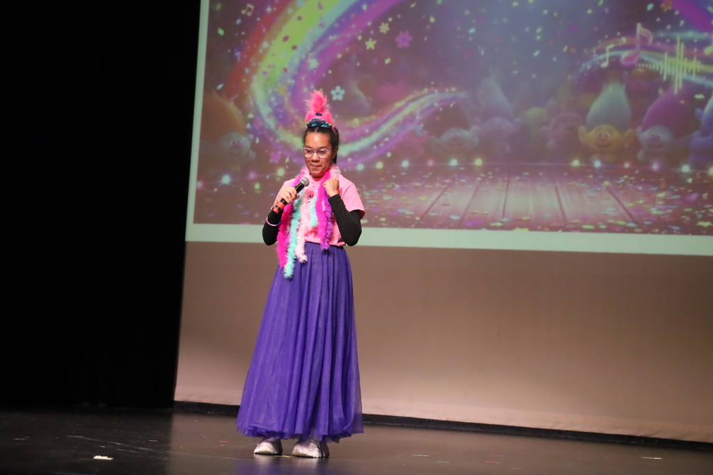 Student in front of the movie Trolls background wearing multiple boas and a troll pink hair piece, singing 