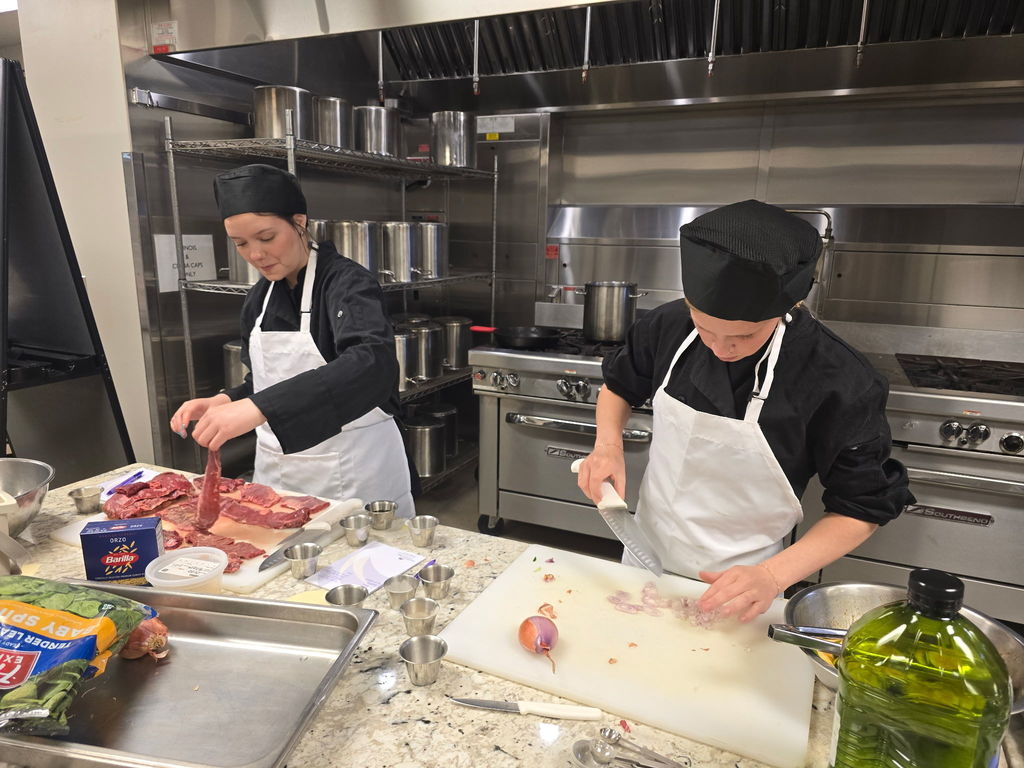 One student handling meat and the other chopping vegetables both in culinary gear