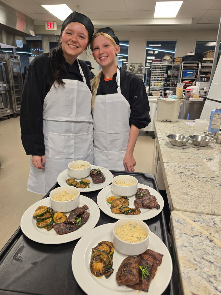 Two students in chef gear with plates in front of them of steak, potatoes and a dressing