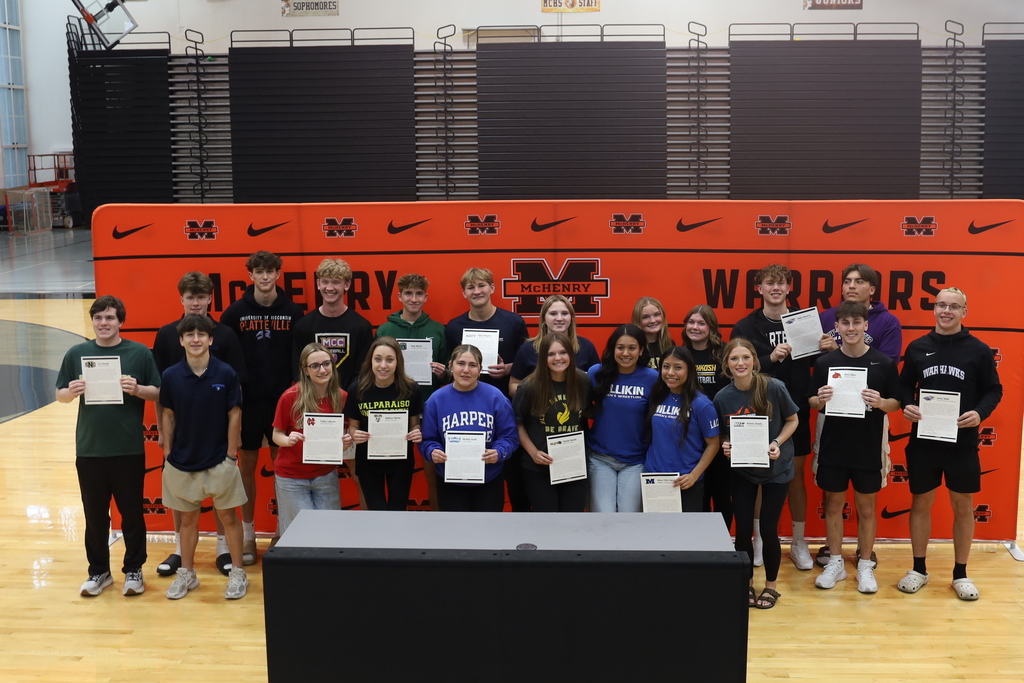 21 students standing in front of McHenry Warriors back drop. Some holding their athletic signing paper