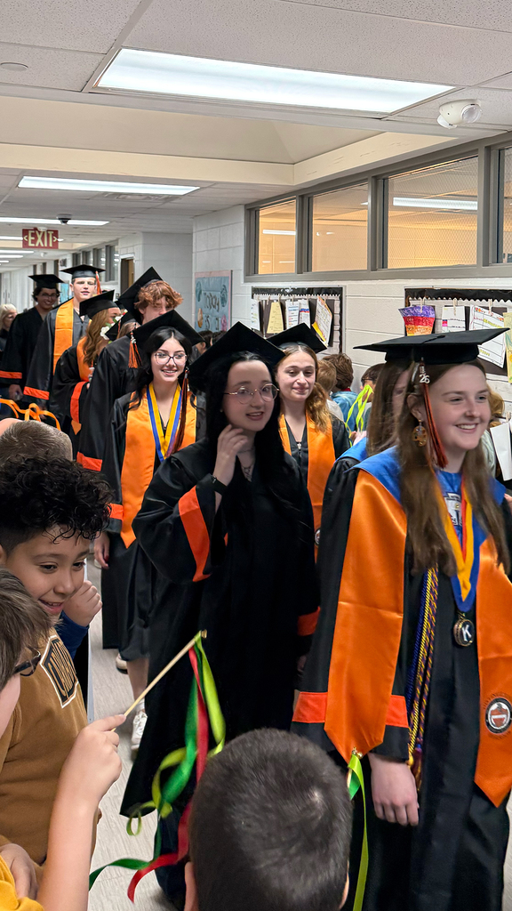 Students walking the halls in caps and gowns with younger students holding ribbons