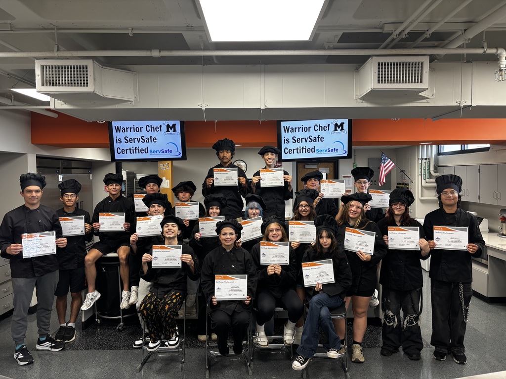 Students in Chef uniforms holding certificates. Warrior is ServSafe Certified on the TV in the background