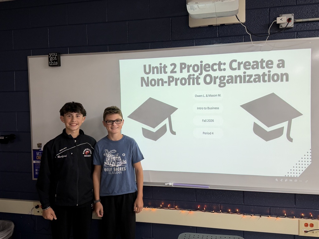 Two students in front of slide that says "unit 2 project create a non-profit organization