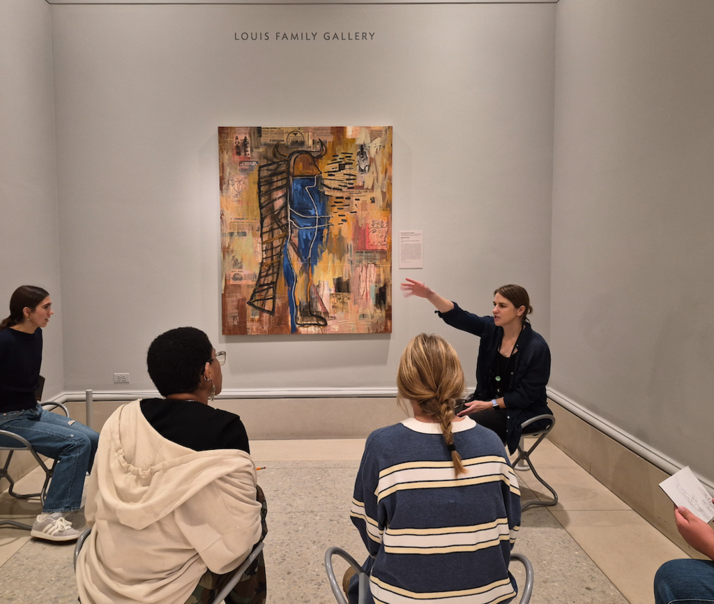 Students sitting in a room looking at an art piece with a tour guide.