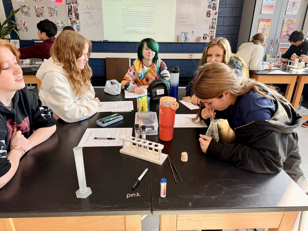 Students at a lab table one exhaling into a straw into the beaker