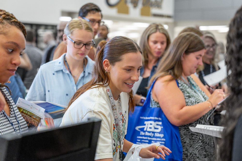 young woman in a crowd smiling as she reaches for item at display table