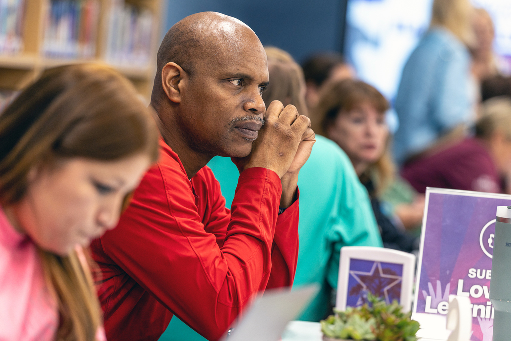 Male teacher seated at table and listening to a colleage