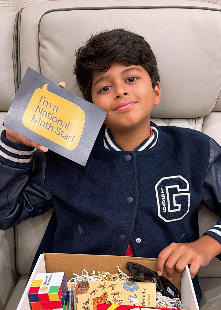 Kiaan sitting in chair with box on his lap and holding card that reads, "I'm a National Math Star!"