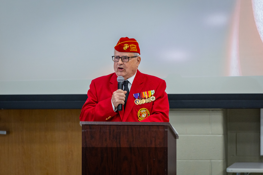 Sgt Douglas Chapman at podium speaking and wearing read uniform of the Marine Corps League