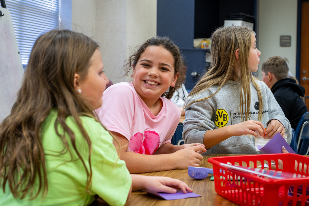 girl at table with peers looking at camera and smiling during activity