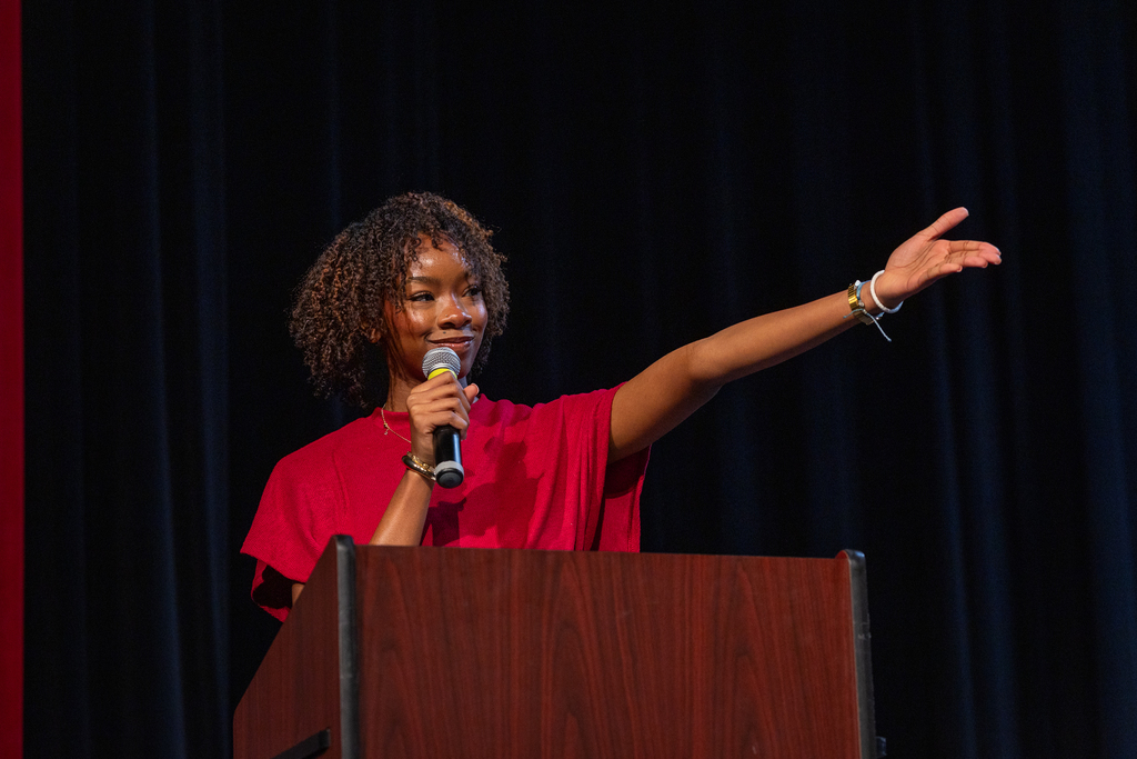 female student at the podium with mic smiling and gesturing toward stage