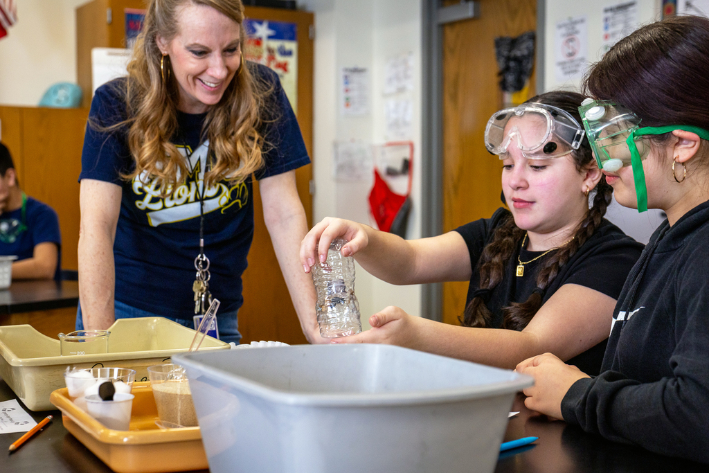 teacher looking on as two female students work on water filtration lab 