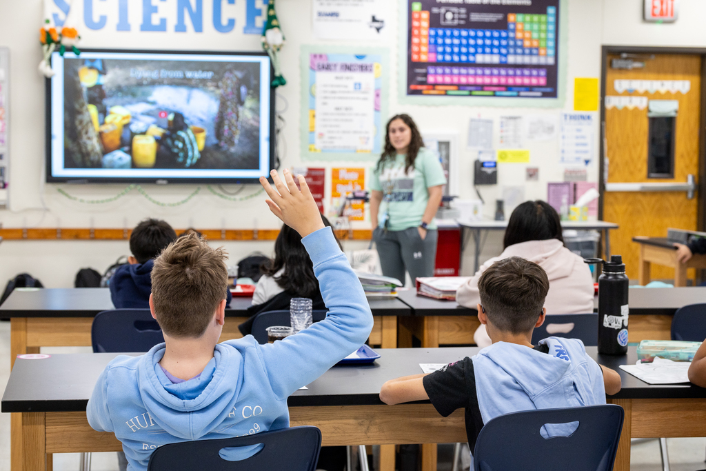 male student raising hand as teacher speaks at front of class