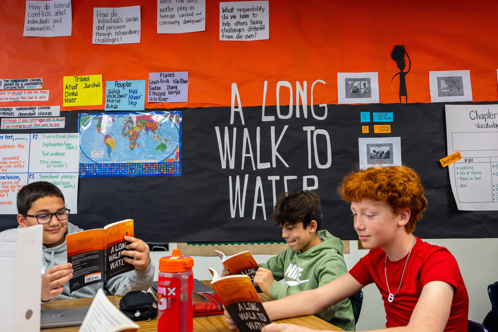 several male students sitting at table reading book A Long Walk to Water with large wall display about the book behind them
