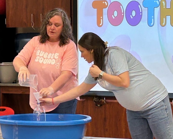 Mrs. McKee and Mrs. Black pour solutions into a bottle to make tiny, bubbled elephant toothpaste.