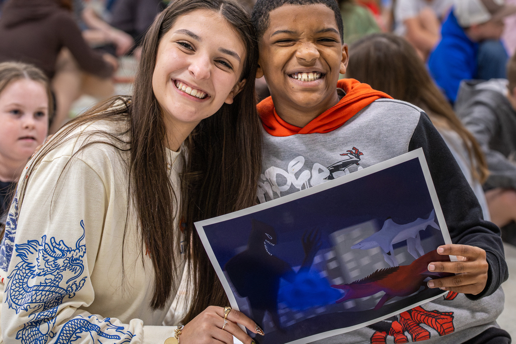 female high school student smiling with male 3rd grader as they hold up the finished art she created for the project