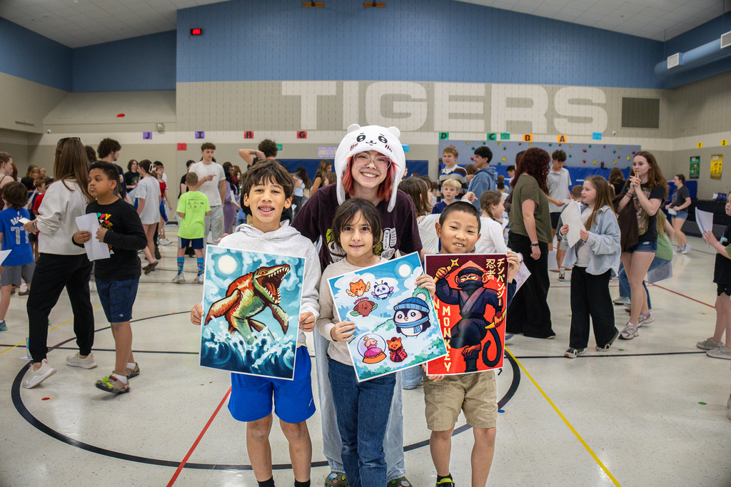 female high school student in fuzzy hat standing behind 3 3rd grade students holding the finished art she completed for the project