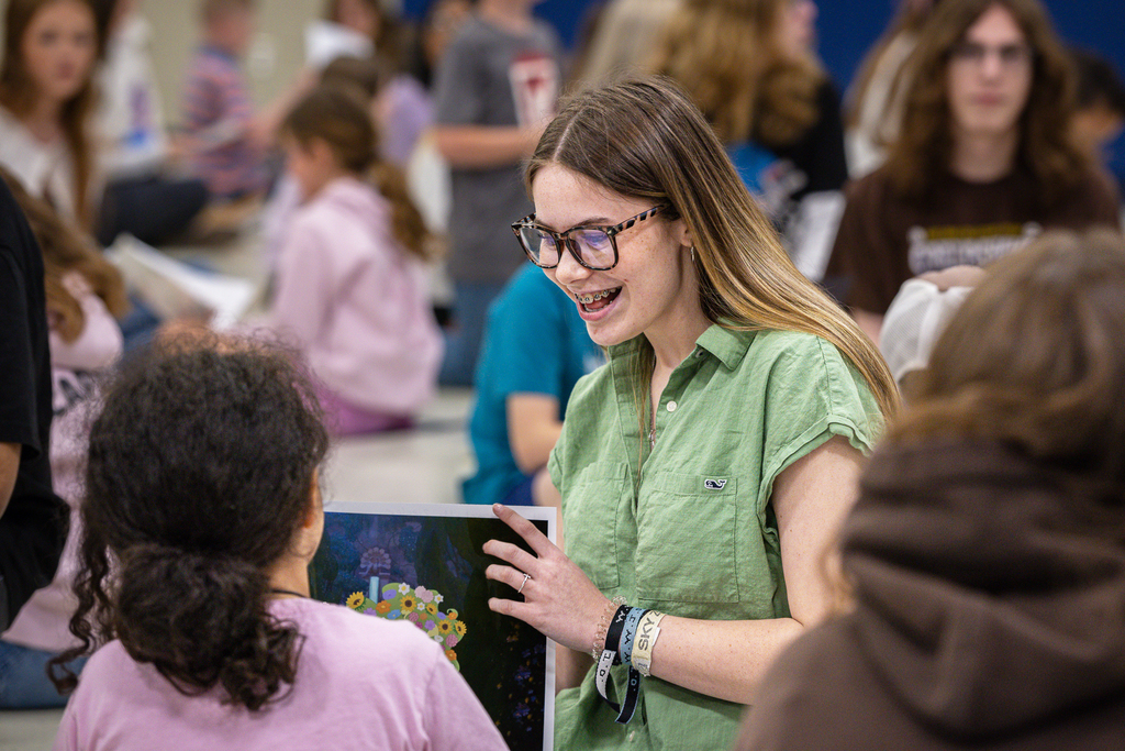 female high school student smiling as she holds up finished art piece for 3rd grade girl to see