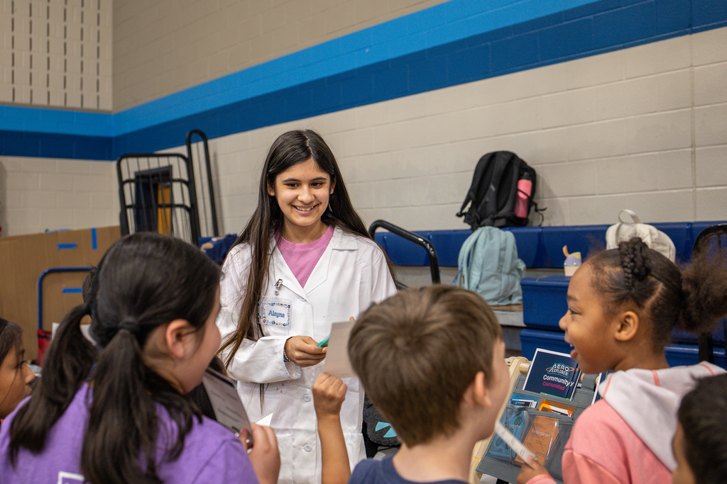 elementary kids gathered around middle school girl in white lab coat