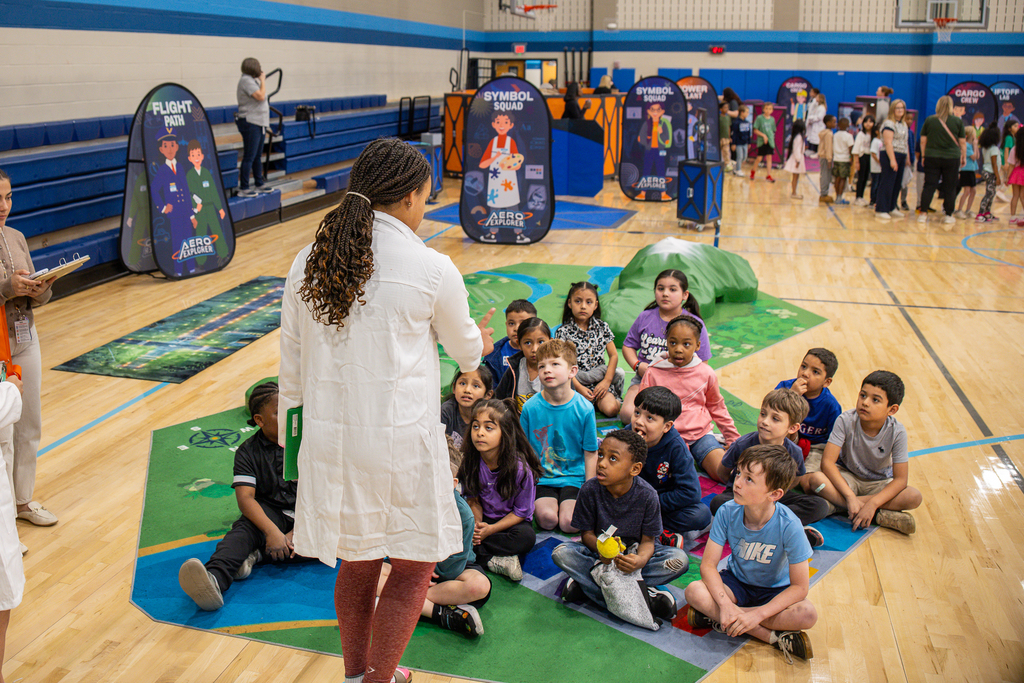 middle school girl in white lab coat standing in front of group of elementary kids talking