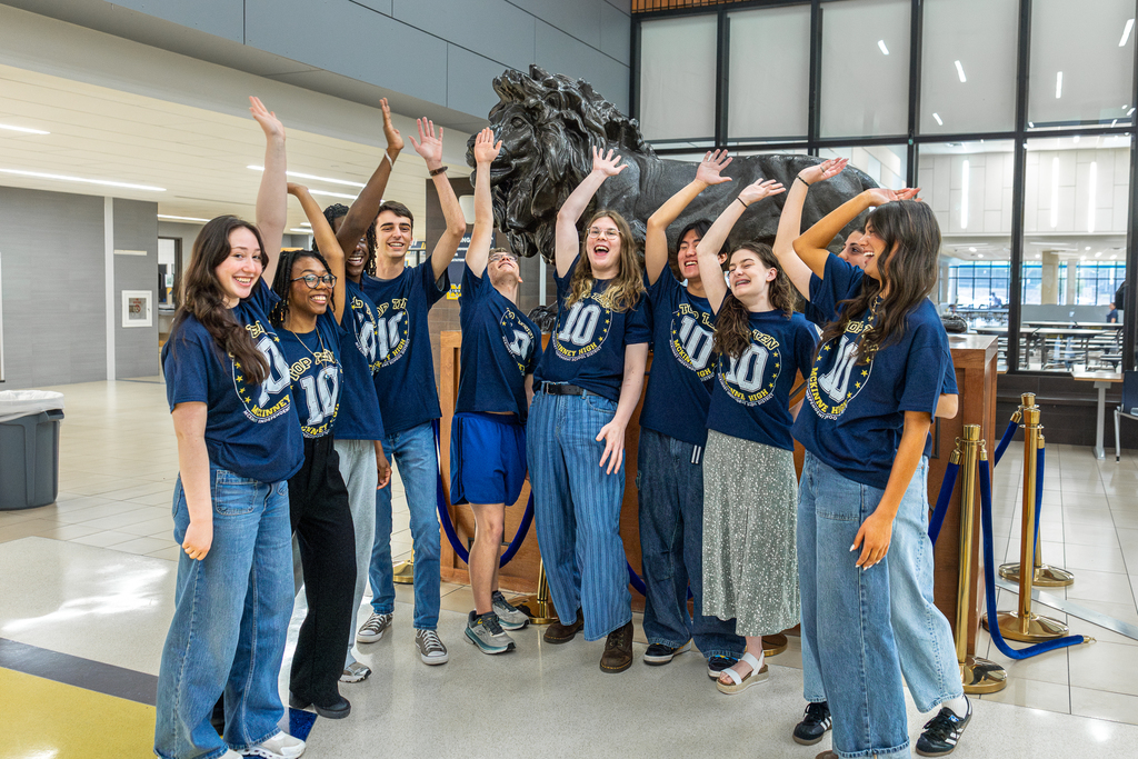 top 10 students in front of lion statue in semicircle with hands in the air cheering