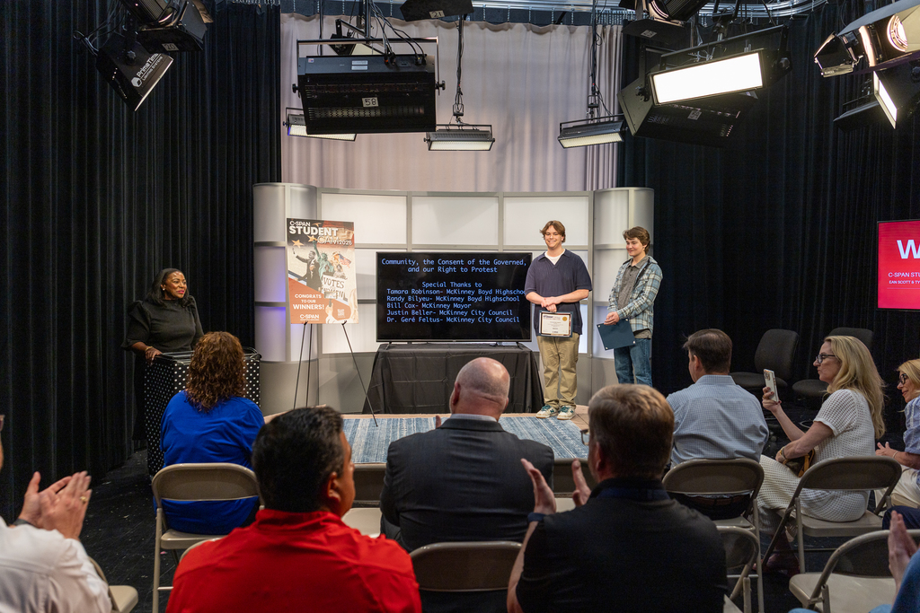 Ean and Tyler on stage as Robin Newton speaks at podium