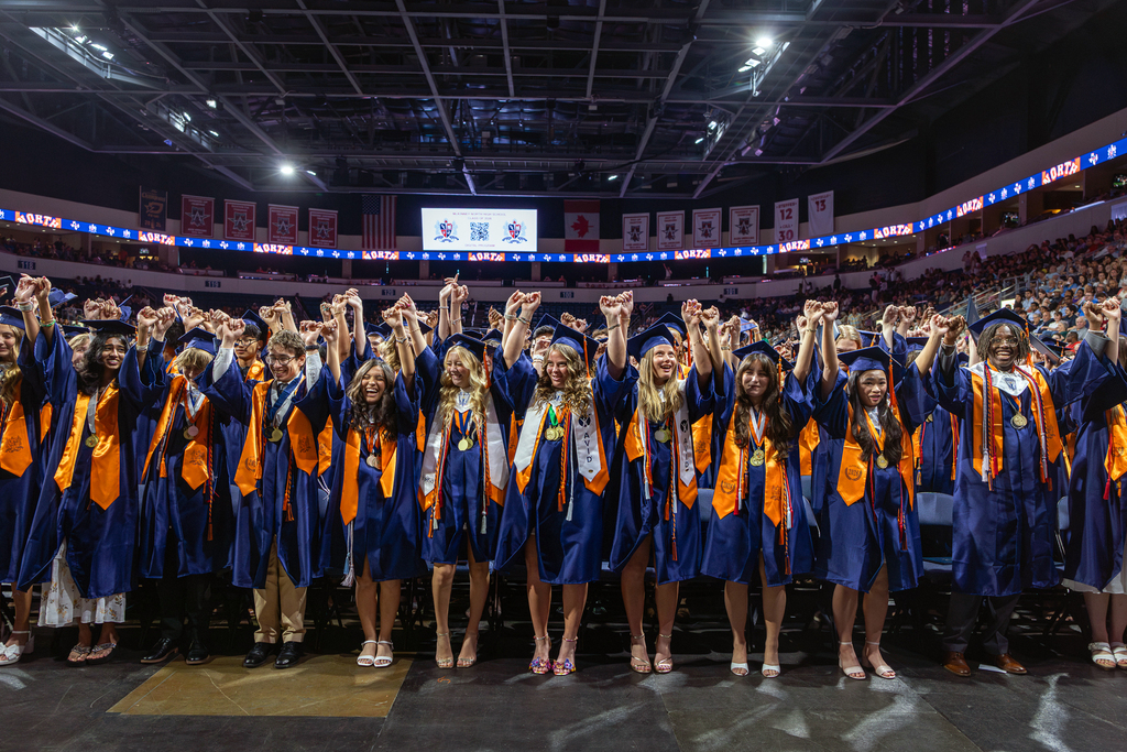 front row of graduates holding hands up and singing the alma mater