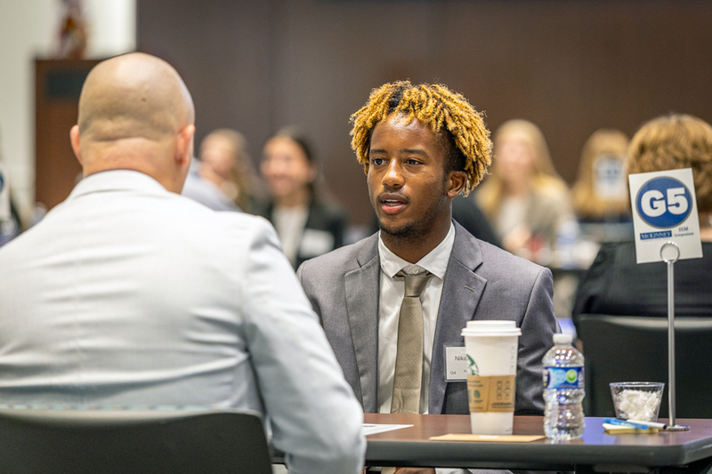 male student talks with an interviewer while seated at a  table