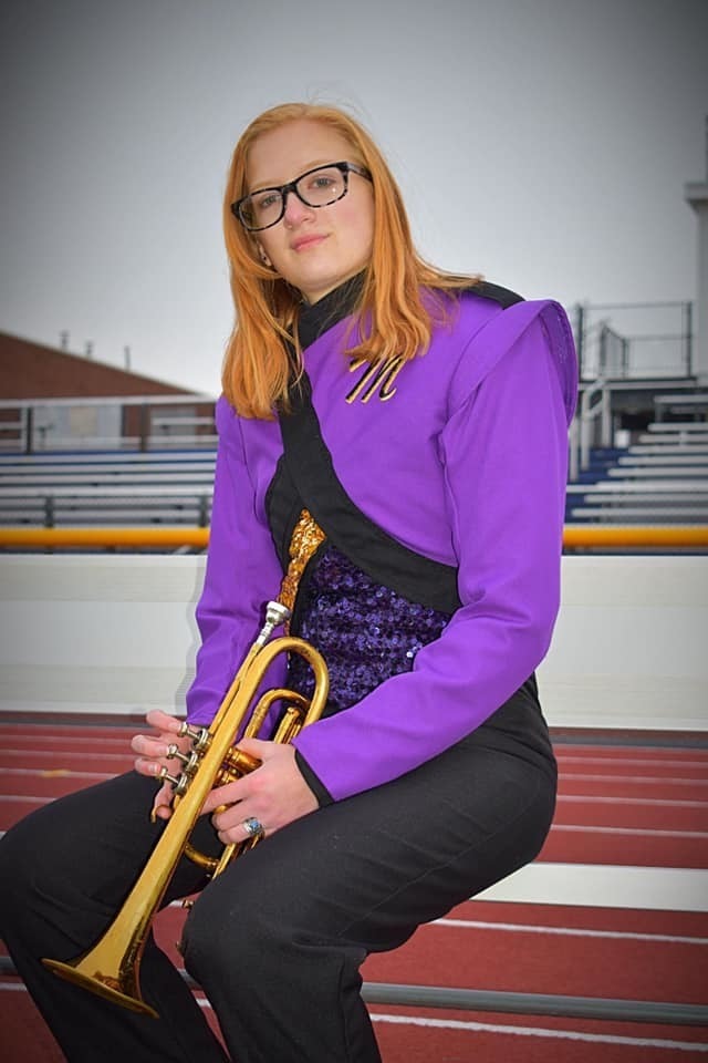 Amber is wearing her purple and black band uniform. Amber is holding her trumpet, and sitting on a bench that is right in front of the track.