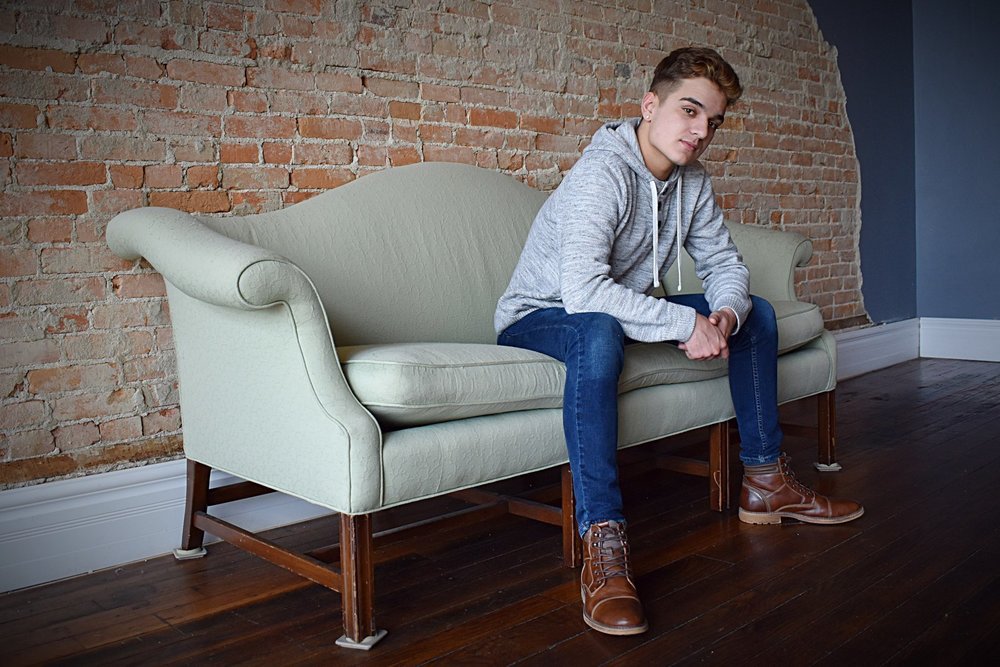 Bailey is wearing a grey sweatshirt with blue jeans and brown boots. Bailey is sitting on a white couch that is sitting in front of brwon brick wall.