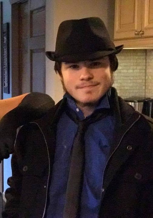 Ben is wearing a blue shirt, black tie, black coat and black hat. Looks like he is standing in a kitchen. I see a light brown cabinet behind him.