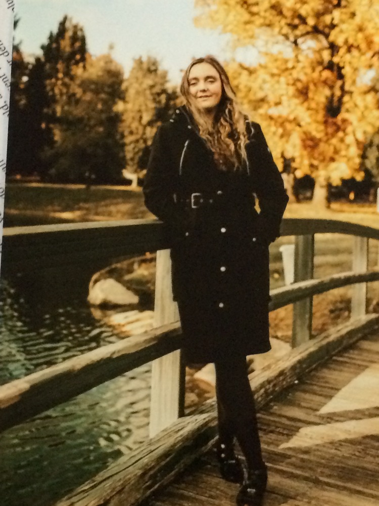Brooke is wearing a black long coat with black boots. She is standing on a bridge, that is over water. It is fall outside. We have tree in the background that the leaves are changing and look yellow. 