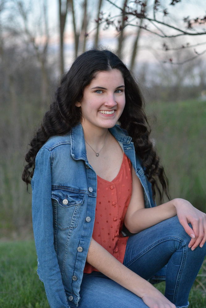 Isabelle is wearing an orange shirt with a blue jean jacket. She is also wearing blue jeans. Isabelle is kneeling down in a green field with trees in the background that have lost their leaves. 