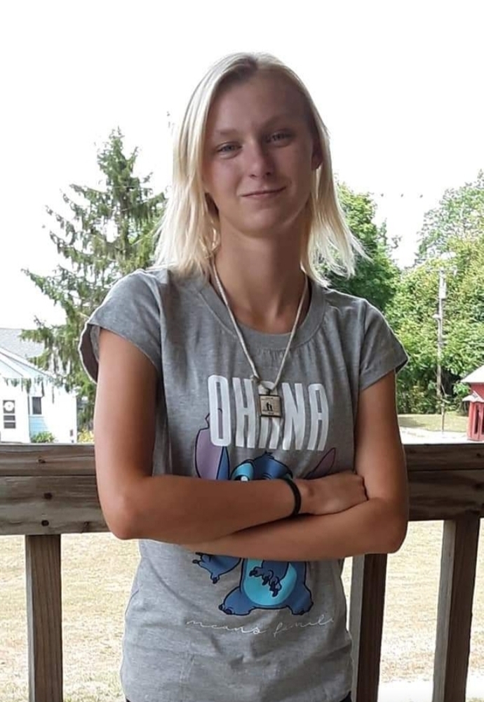 Anna is wearing a grey tshirt with white writing. cant read it due to her wearing a neckles that hangs down. She is standing in front of a brown railing.