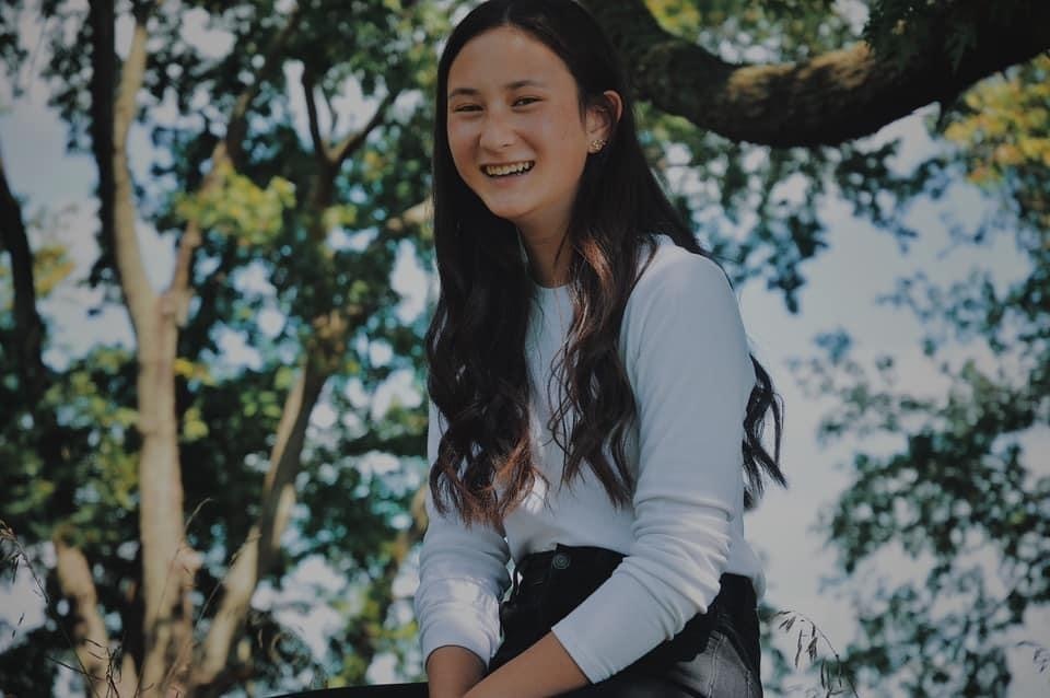 Gloria is wearing a white long sleve shirt and black jeans. Gloria is sitting in a tree. The trees are in full bloom of green and it is nice and sunny out.